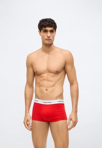 Enkeltmann uten skjorte med kort krøllete hår iført røde Calvin Klein boxershorts som står mot en ensfarget lys bakgrunn.