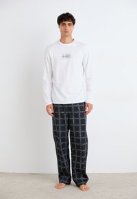 Cămașă albă cu mâneci lungi și logo imprimat negru; pantaloni de pijama negri cu un model în grilaj alb; modelul stă desculț pe un covor bej.