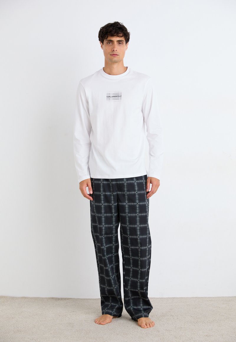 Cămașă albă cu mâneci lungi și logo imprimat negru; pantaloni de pijama negri cu un model în grilaj alb; modelul stă desculț pe un covor bej.