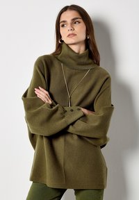 Pull à col roulé oversize de couleur vert olive en tissu doux, avec des manches longues à détail boutonné et une coupe décontractée.