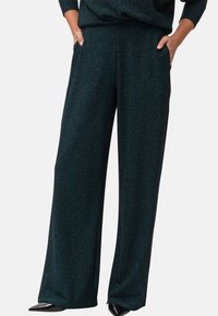Femme portant un pantalon large vert foncé avec un léger éclat et les mains dans les poches, assorti à des chaussures noires pointues.