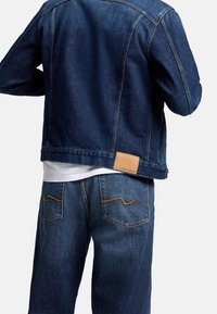Giacca in denim scuro con taglio dritto e cuciture visibili, abbinata a jeans in denim scuro. Presenta una toppa in pelle marrone sul retro.