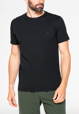 Uomo con la barba che indossa una semplice t-shirt nera a maniche corte con scollo rotondo e pantaloni verde oliva, leggermente rivolto verso sinistra su sfondo bianco.