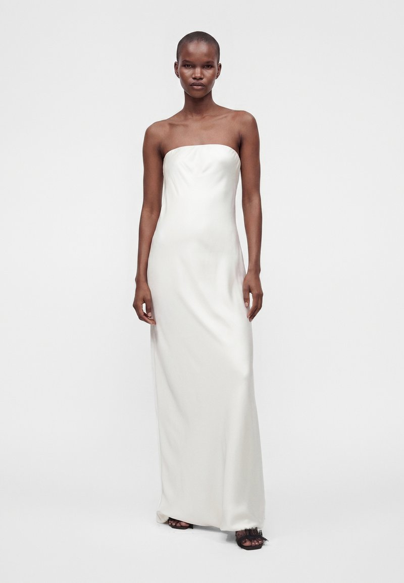 Femme portant une robe blanche longue bustier avec des talons ouverts noirs, debout devant un fond clair uni.