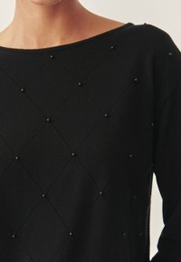 Schwarzer Pullover mit Rautenmuster und kleinen schwarzen Perlen an den Kreuzungspunkten, getragen von einer Person mit sichtbarem Hals und Schulter.