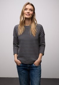 Femme aux longs cheveux blonds portant un pull bleu marine à motifs et un jean bleu, debout les mains dans les poches devant un fond gris uni.