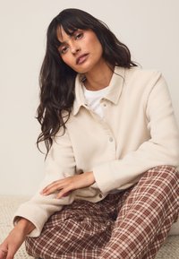 Giacca in pile color crema con chiusura a bottoni a pressione, design con colletto e maniche lunghe, abbinata a pantaloni a quadri in tonalità terrose.