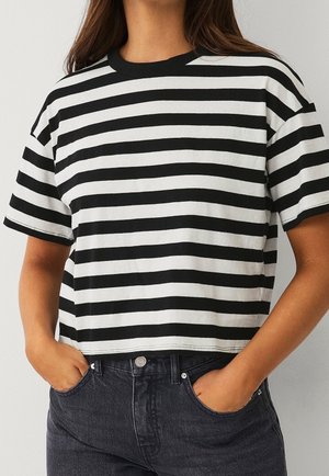 Femme portant un crop top rayé horizontal noir et blanc à manches courtes avec col noir, mains dans les poches d'un jean foncé taille haute.