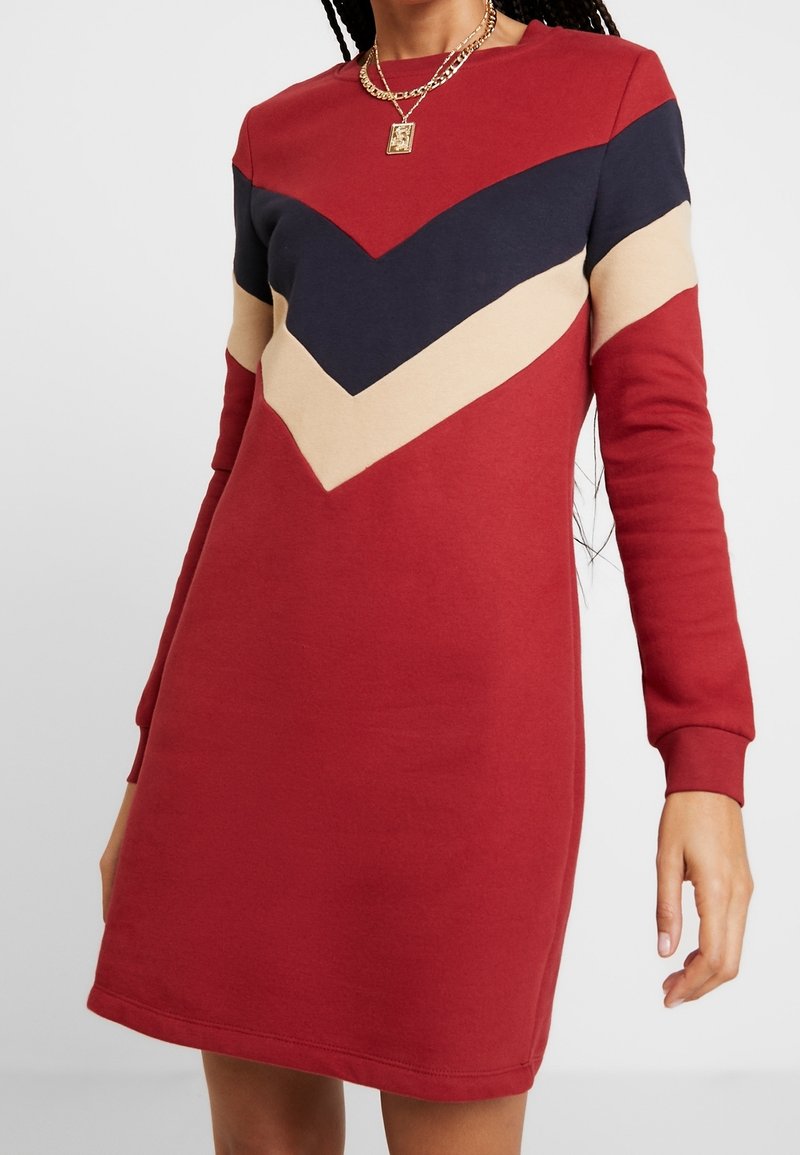 Femme portant une robe rouge à manches longues avec des rayures chevrons bleu marine et beige sur la poitrine et le haut des bras, accessoirisée avec des colliers dorés.