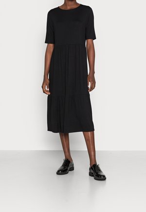 Kvinde iført en sort midi-kjole med korte ærmer og skinnende sorte snøresko, stående foran en ensfarvet lys baggrund.