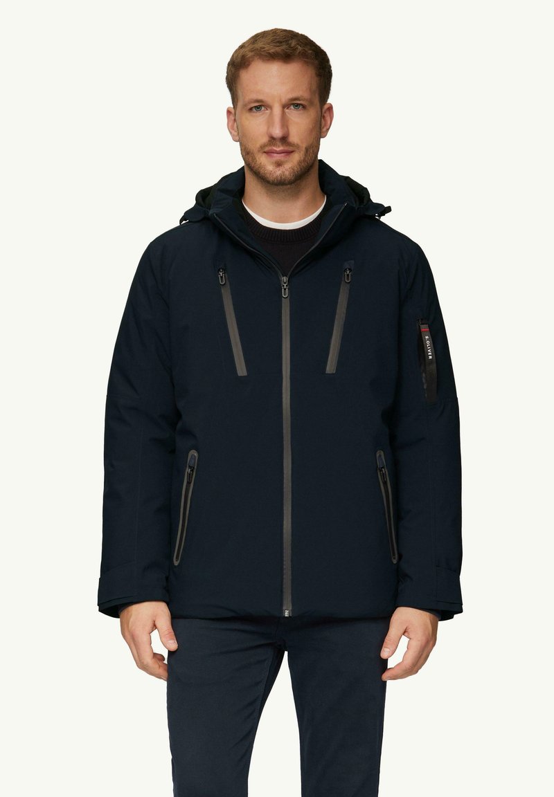 Veste étanche bleu marine foncé avec fermeture éclair à l'avant, capuche réglable et plusieurs poches, avec des zips et des accents gris contrastants.