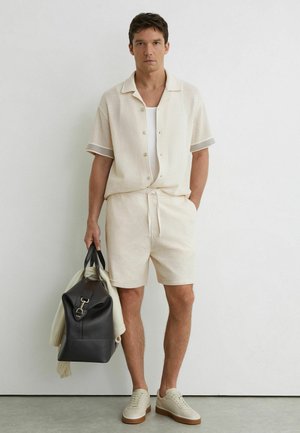 Bărbat purtând o cămașă bej cu mânecă scurtă și pantaloni scurți asortați, ținând o geantă de voiaj din piele neagră, peste care este așezat un pulover crem.