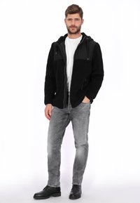 Chaqueta polar negra con cremallera, tejido texturizado, acentos de nylon negro, capucha y bolsillos delanteros. Combinada con vaqueros gris claro y zapatos negros.