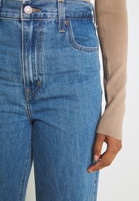 Gros plan d'une personne portant un jean bleu taille haute et un haut beige à manches longues, main détendue le long du corps sur fond clair.