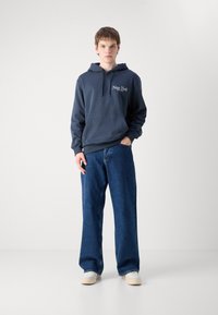 Marineblå hettegensar med "New York" tekst, frontlomme, kombinert med løse denimjeans og lyse sneakers. Modellen står mot en ensfarget bakgrunn.
