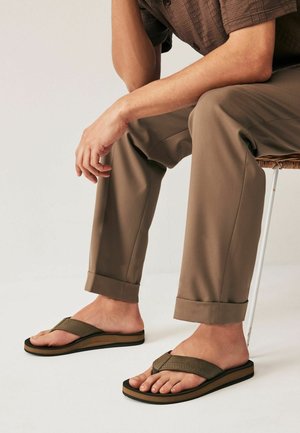 Sandali infradito marroni con una fascia ampia, parte superiore testurizzata e suola spessa. Indossati con pantaloni khaki, seduto su una sedia intrecciata.