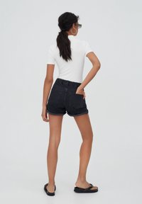 Shorts in denim nero con cintura elastica, risvolti e due tasche posteriori, abbinati a una maglietta bianca aderente e sandali neri.