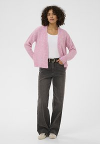Cardigan rosa peloso con davanti aperto, indossato sopra una maglietta bianca, abbinato a jeans neri a vita alta e sneakers di colore chiaro.
