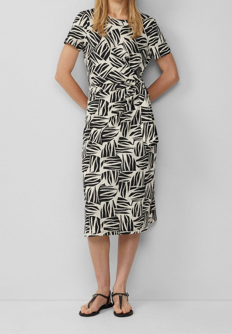 Femme portant une robe noire et blanche à motif feuilles longueur genoux, manches courtes et sandales plates noires, debout devant un mur uni.