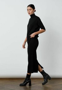 Sort turtleneck-top parret med en sort midi-nederdel med sideslids. Modellen har sorte hælede ankelstøvler på. Glat tekstur, tætsiddende design.