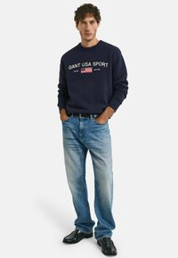 Navy sweatshirt with "GANT USA SPORT" print and flag detail, paired with light blue jeans and black shoes, featuring a relaxed fit and cuffs.