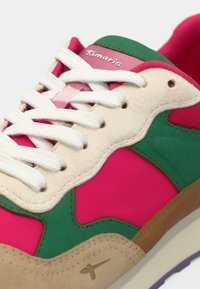 Gros plan d'une chaussure de sport avec des lacets blancs, du daim beige, des sections en textile vert et rose, et des accents marron sur la semelle.