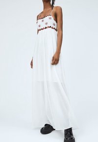 Weißes Maxikleid mit tailliertem Oberteil, buntem floralen Stickerei, dünnen Trägern und einem fließenden, transparenten Rock. Getragen mit schwarzen klobigen Schuhen.