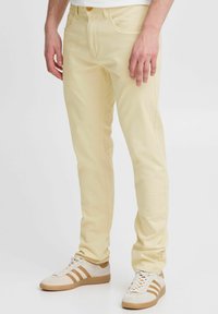 Pantalones de mezclilla amarillo claro con corte recto, bolsillos frontales y cierre de botón, combinados con zapatillas deportivas beige con detalles en marrón.