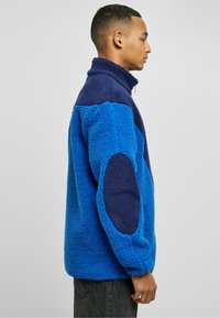 Blue and navy fleece pullover with a high collar, textured fabric, and elbow patches, featuring a zipper closure and relaxed fit.