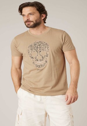 Mann mit Bart, trägt ein beiges T-Shirt mit floralem Totenkopf-Design und weiße Cargo-Shorts, steht mit einer Hand in der Tasche.