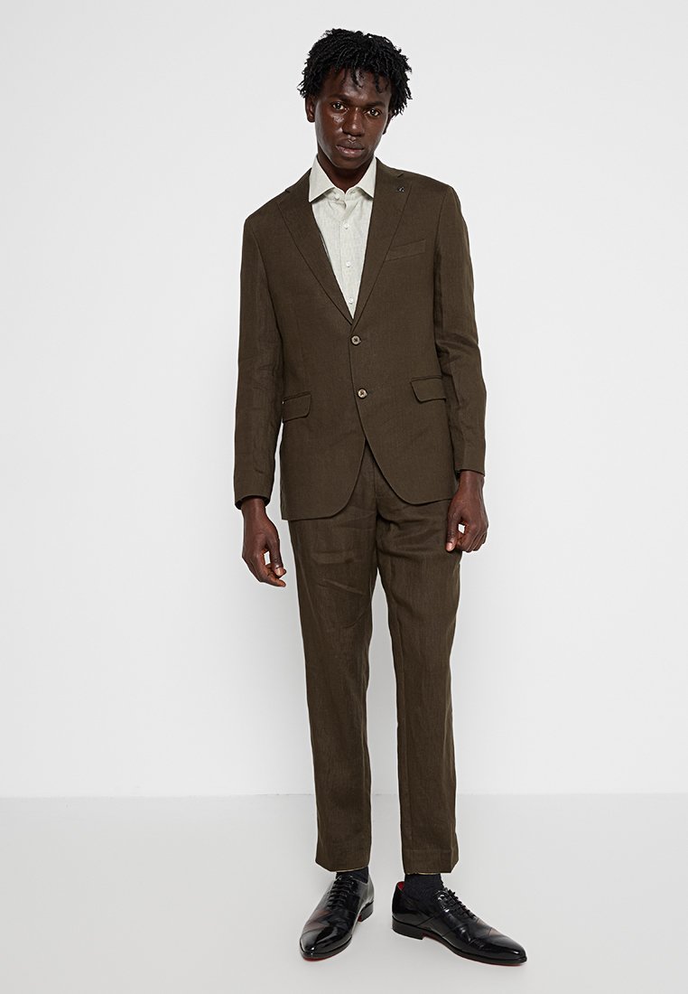 Dunkelbrauner Leinenanzug mit einem Zwei-Knopf-Sakko, kombiniert mit einem hellen gemusterten Hemd. Schwarze, polierte Schuhe vervollständigen das formelle Outfit.
