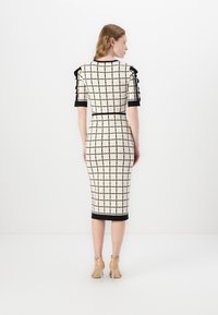 Rochie midi ajustată, de culoare crem, cu un model în carouri negre, mâneci scurte cu accente volănașe și tiv solid negru. Tocuri transparente vizibile.