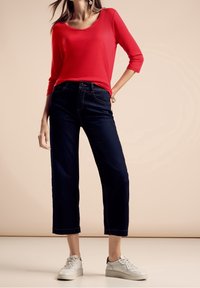 Rotes Langarmshirt kombiniert mit dunkelblauen, kurzen Jeans. Weiße Sneakers mit grauen Akzenten. Glatter Stoff und taillierte Passform.