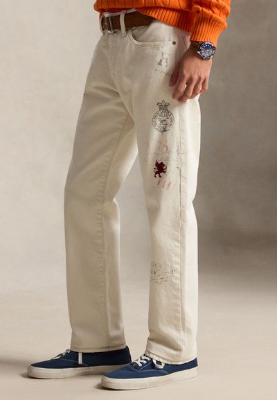Person iført off-white jeans med trykte logoer, blå joggesko, brun belte, oransje genser og en klokke som står mot en nøytral bakgrunn.