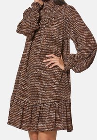 Braun gemustertes Kleid mit langen Ärmeln, hohem Kragen und fransigem Saum. Hergestellt aus strukturiertem Stoff mit einer Mischung aus hellen und dunklen Farbtönen.