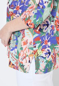 Blusa a fiori nei colori verde, rosa, blu e arancione, con maniche corte, spacchi laterali e un tessuto leggero e testo.