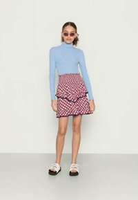Gola alta azul claro, saia mini em camadas com estampa gráfica nas cores vermelho, branco e azul, combinada com sandálias brancas de solas pretas.