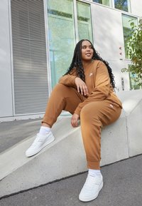 Brun matching hettegenser og bukser med ribbestrikkede mansjetter, paret med hvite sneakers og ankelsokker, sittende på en betongkant.