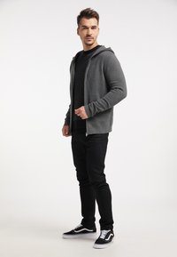 Grijze rits-hoodie over een zwarte gestructureerde trui, gecombineerd met zwarte slim-fit jeans en zwarte sneakers met witte accenten.