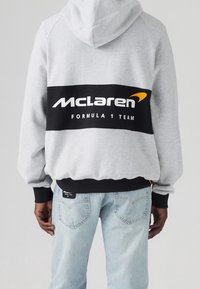 Szary hoodie z czarnym pasem z logo "Zespół McLaren Formuły 1", regulowany kaptur, ściągacze u rękawów, w połączeniu z niebieskimi dżinsami.