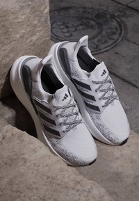 Chaussures de course Adidas UltraBoost blanches et grises avec des lacets à motifs placées sur des marches en pierre à côté d'une plaque d'égout.