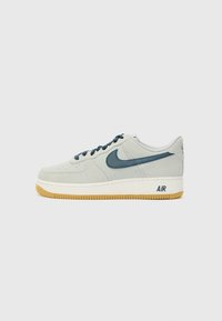 Vaaleanharmaa mokkanahkainen tennari, jossa on tummansininen Nike-swoosh, pyöreä kärki, litteät nauhat, valkoinen välikenkä ja kumipohja. "AIR" painettuna sivussa.