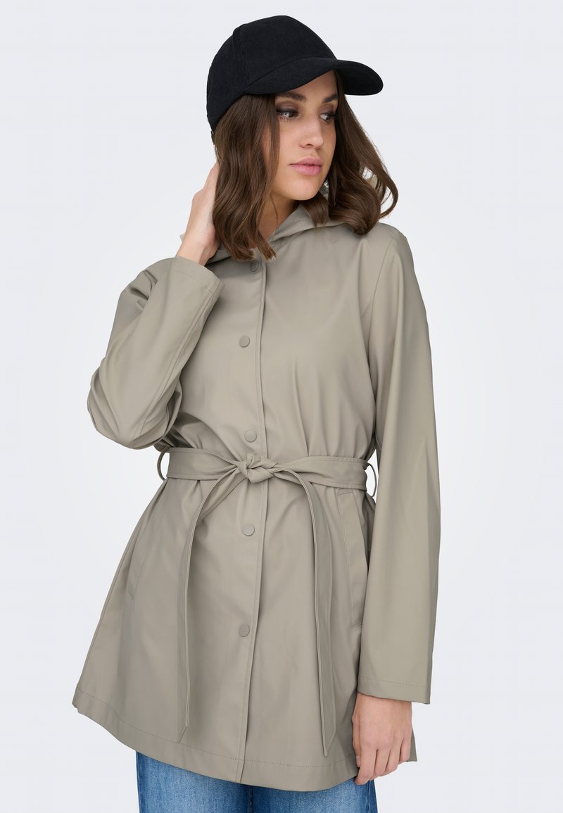 Mujer con impermeable gris claro con cinturón y gorra negra, con cabello suelto ondulado y castaño, mirando de lado con una mano tocándose el cuello.