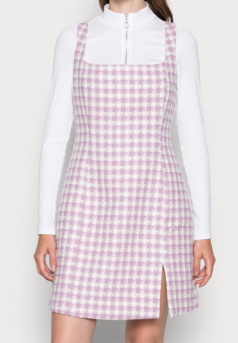Femme portant une robe sans manches à carreaux rose et blanc avec une fente à l’avant, par-dessus un top blanc à manches longues avec un col zippé.