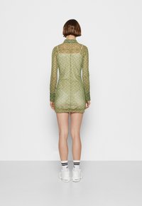 Vestido ajustado de mangas longas feito de um tecido verde transparente e com padrão, apresentando um colarinho e um design até a metade da coxa, combinado com ténis brancos.