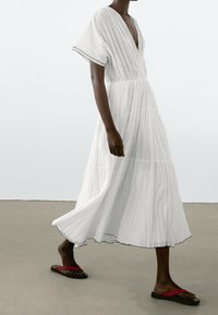 Personne à la peau foncée marchant dans une robe blanche fluide à col en V avec manches courtes et sandales à lanières rouges sur un sol neutre.