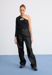Svart topp med én skulder og rund logo, kombinert med høyt liv mørke denimjeans. Modellen har på seg svarte sportsko. Enkel, teksturert bakgrunn.