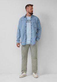 Helles blaues Denim-Hemd über einem hellblauen Grafik-T-Shirt, kombiniert mit hellgrünen Hosen und beigen Sneakers. Minimalistisches Design und lockere Passform.