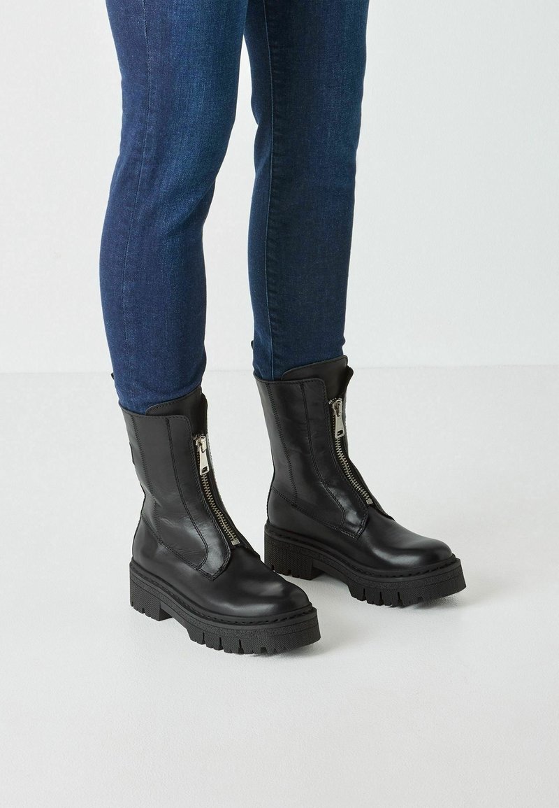 Bottes pour femmes en cuir noir à talons épais et fermetures éclair à l'avant, portées avec un jean skinny bleu foncé sur fond blanc.
