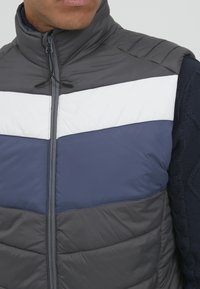 Vesta căptușită, cu guler înalt, dintr-un material gri cu dungi în zigzag albe și bleumarin și închidere cu fermoar. Detaliu cu mâneci din tricot texturat vizibil.